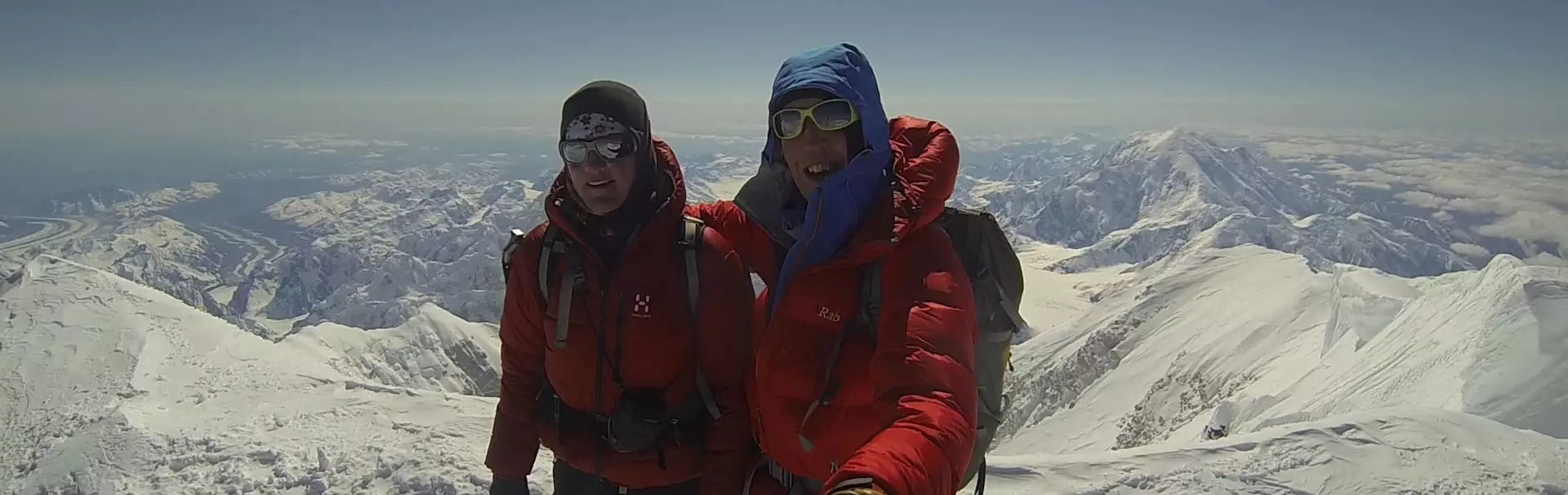 Een persoon in een rode jas staat triomfantelijk op een besneeuwde bergtop met uitgestrekte armen en een ijsbijl in de hand. Een rugzak en klimuitrusting liggen naast hem in de sneeuw, onder een strak